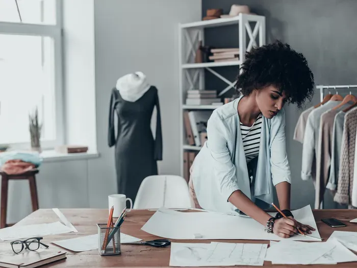 Young fashion designer sketching at a table surrounded by clothing racks, patterns, and design materials